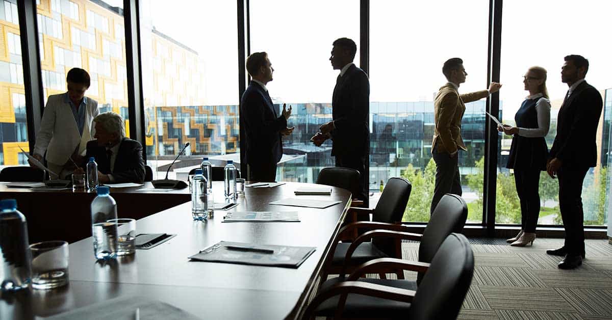 Business meeting in a modern office with large windows. Six people are engaged in discussions or looking at documents around a table with bottles and papers.