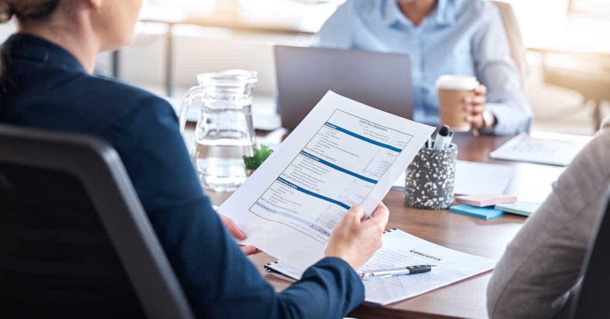 Person holding a financial report in an office meeting. Desk has a laptop, coffee cup, water jug, potted plant, pen, and notebook. Two blurred people in the background.