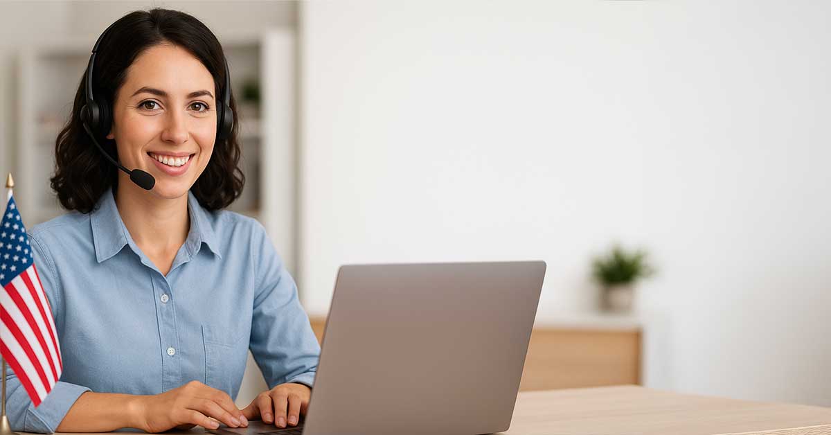 A woman wearing a headset sits at a desk with a laptop and a small American flag, smiling at the camera in a bright office setting.