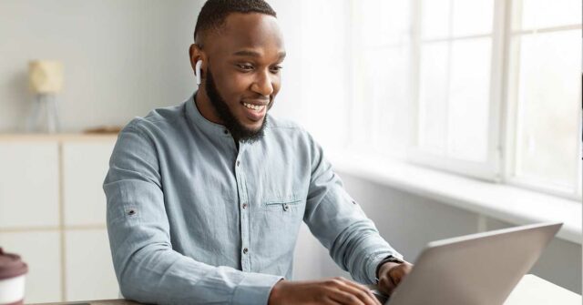 A man wearing earbuds and a light blue shirt sits at a desk, smiling while typing on his laptop near a bright window during Open Enrollment.