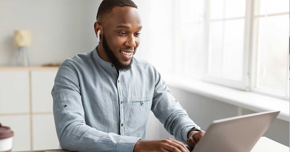 A man wearing earbuds and a light blue shirt sits at a desk, smiling while typing on his laptop near a bright window during Open Enrollment.