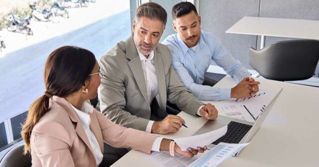 Three people in business attire sit at a desk with documents and a laptop, discussing paperwork and DEV strategies in a modern office setting.