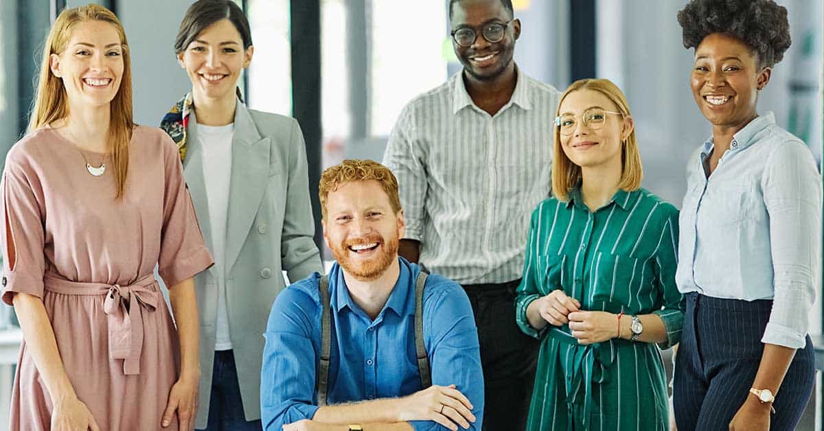Six people stand and smile together in a brightly lit office setting, wearing business casual attire and celebrating the benefits of teamwork.