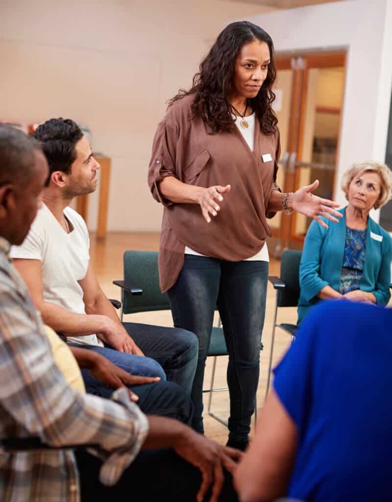 A woman stands and speaks to a seated group of adults in a discussion circle in a brightly lit room.
