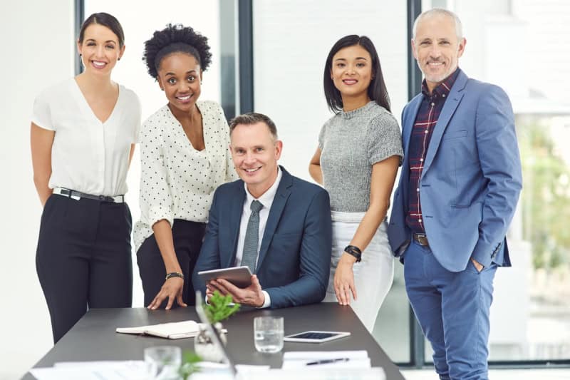 Five professionally dressed people pose together in an office setting, with one man seated at a desk holding a tablet and four colleagues standing behind him, smiling at the camera.