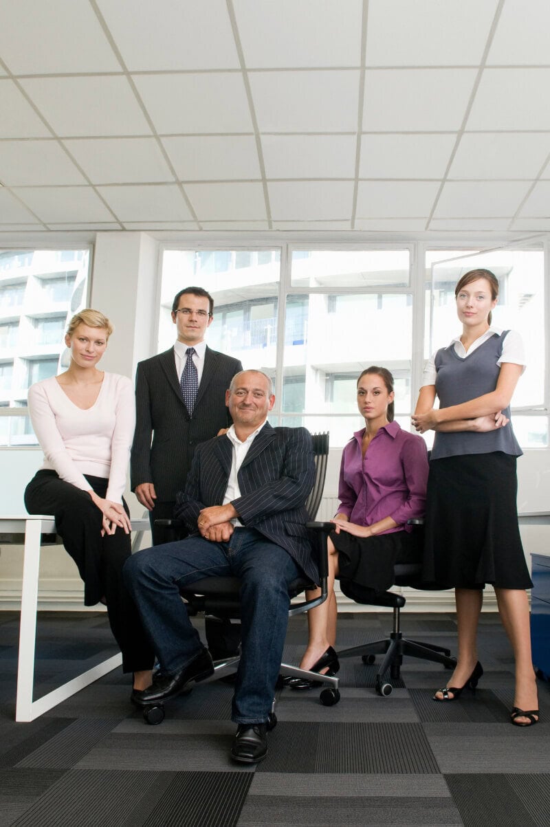 Five professionally dressed people pose together in an office with large windows and city views, with one man seated in the center and the others standing or sitting around him.