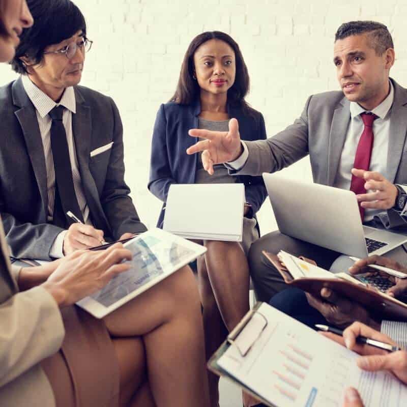 A group of business professionals in suits sit in a meeting, discussing documents related to Benefit Administration Services, with laptops and charts in hand.