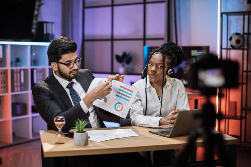 Two people sit at a desk; one person shows charts on paper to the other while speaking, with a laptop, hourglass, and potted plant nearby, and a camera recording them.