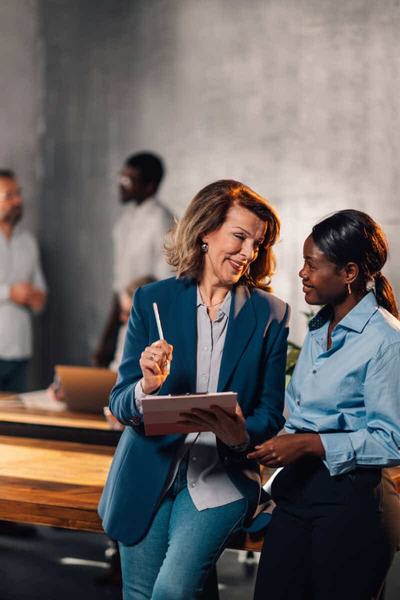 Two women in business attire stand by a desk, conversing and looking at a tablet, while three colleagues interact in the background of an office setting.