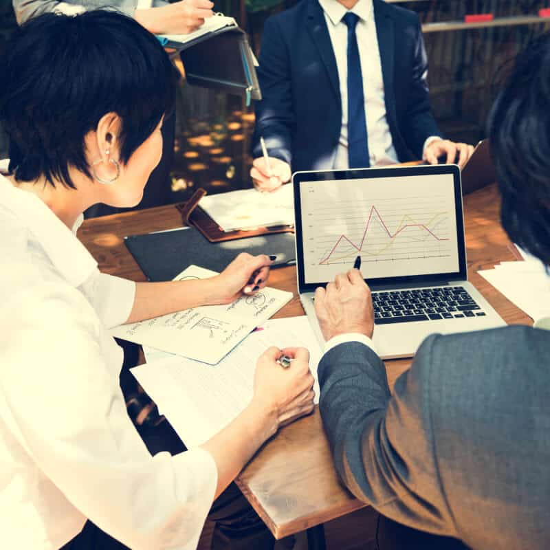 Four professionals in business attire discuss data, examining a laptop with a line graph and reviewing documents at a meeting table.