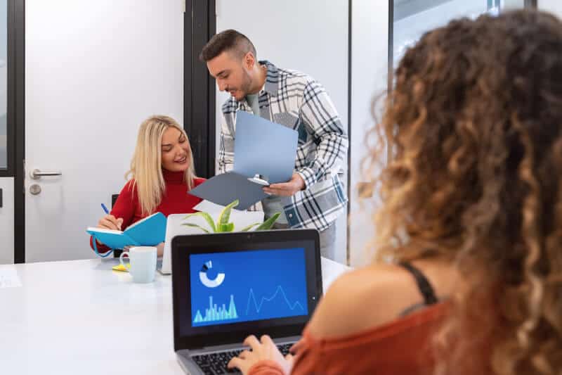 Three people in a modern office; one woman works on a laptop showing charts, while two colleagues discuss documents at the table.