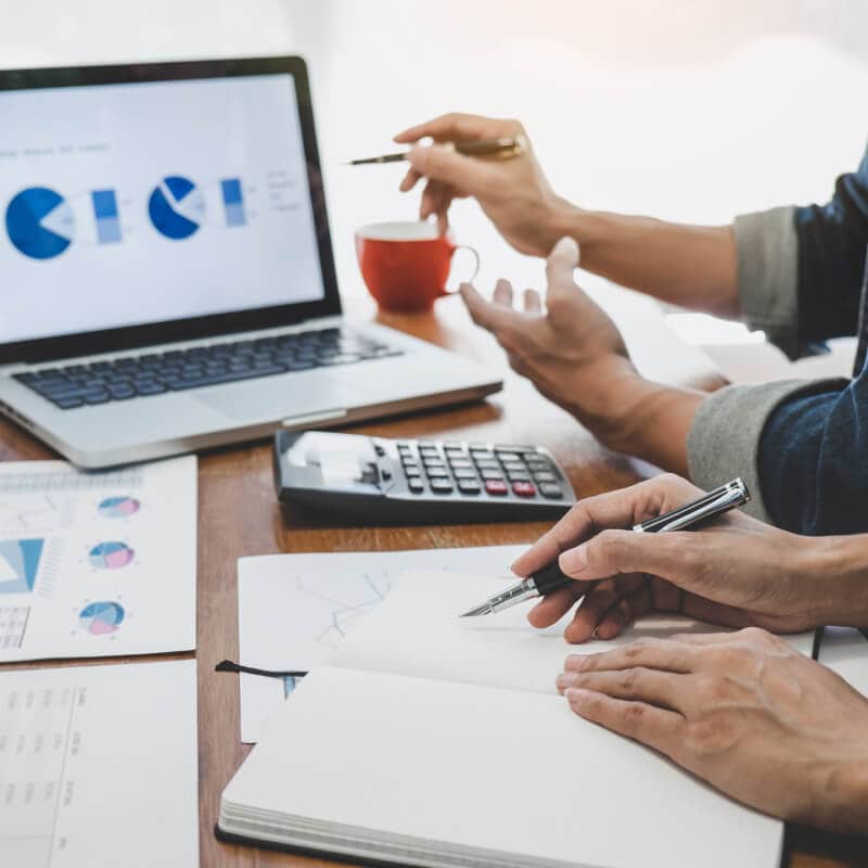 Two people discuss financial charts on a laptop and papers, with a calculator and notebook on the table.