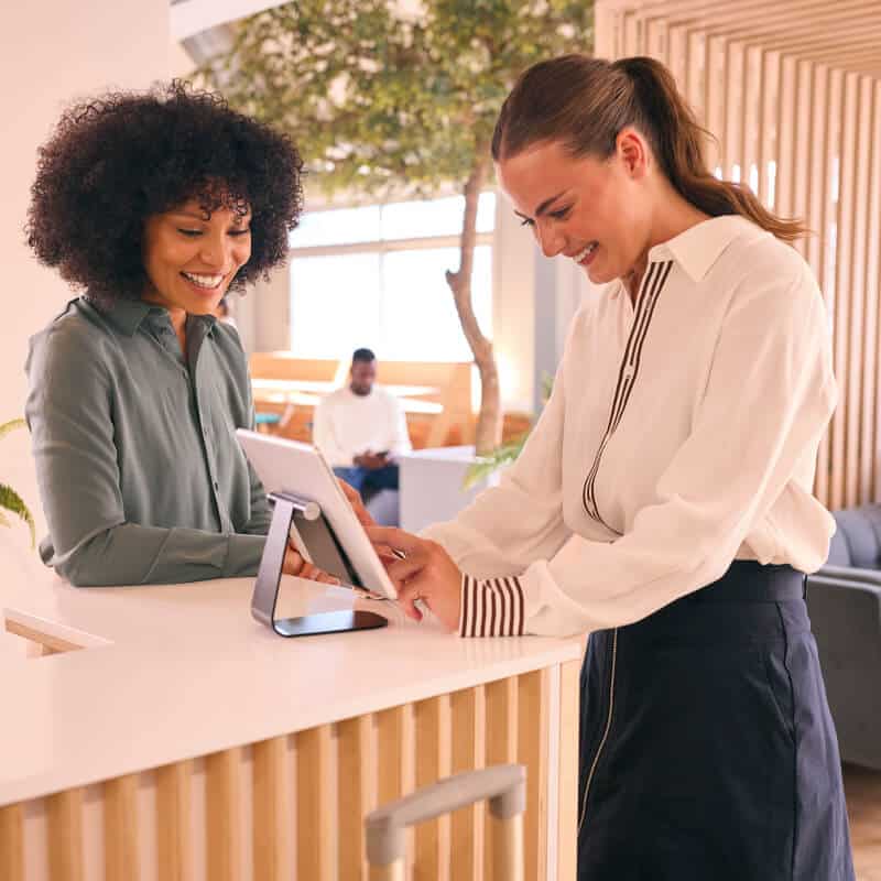 Two women stand at a reception desk, one using a tablet while the other looks on and smiles. A man sits in the background in a modern, well-lit office space.