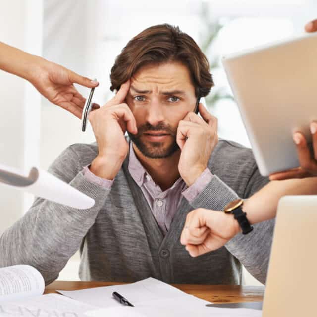 A man looks stressed at his desk while multiple hands offer him a phone, pen, papers, a tablet, and gesture at a watch, suggesting he is overwhelmed with tasks.