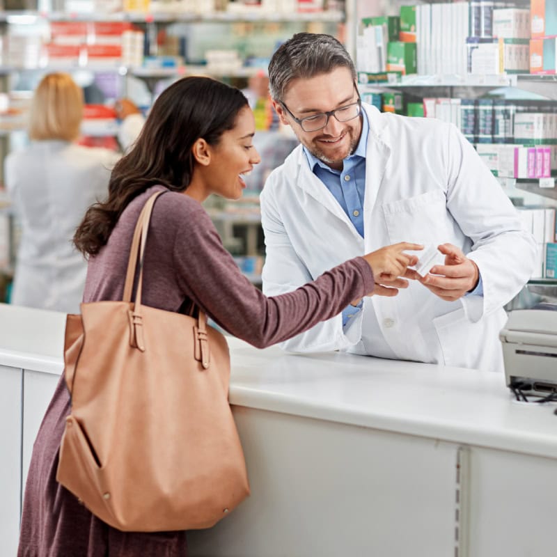 A pharmacist in a white coat explains a medication bottle to a customer at the pharmacy counter. Shelves with medicine are visible in the background.