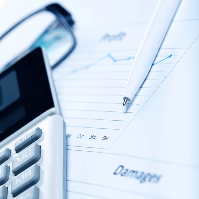 A close-up of a calculator, pen, and eyeglasses on top of documents showing a profit graph and the word "Damages.