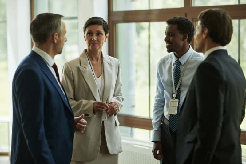 Four professionally dressed people stand in a group having a conversation indoors near large windows.