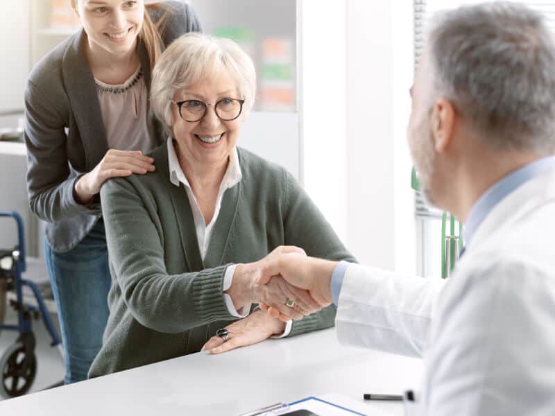 An older woman sitting at a table smiles and shakes hands with a doctor, while a younger woman stands beside her, smiling.