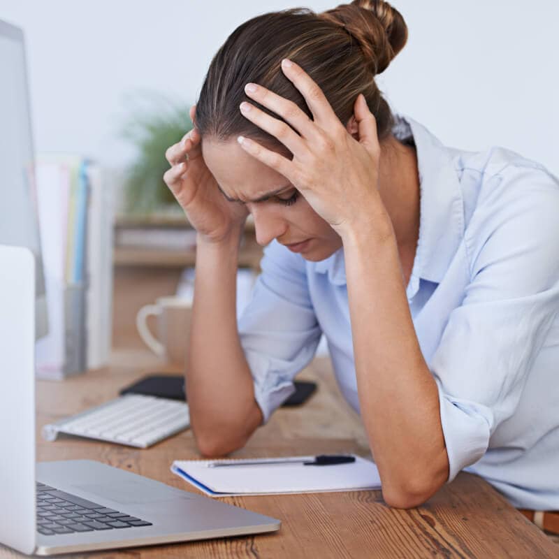 Woman sitting at desk with hands on her head, appearing stressed or frustrated, next to a laptop, notepad, and pen.