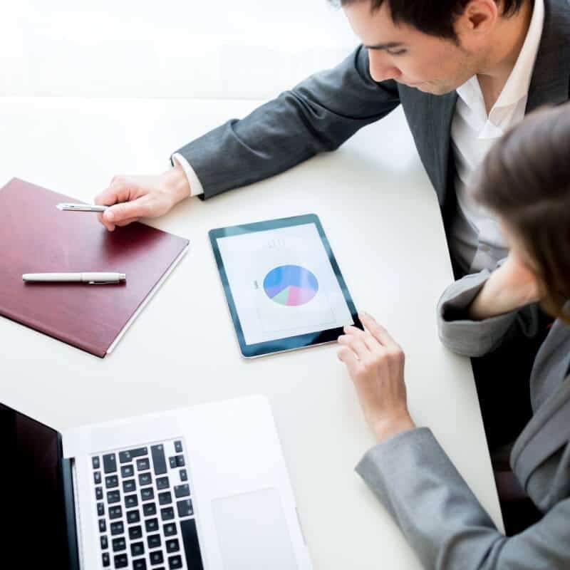 Two people in business attire review a tablet displaying a pie chart, discussing Carrier Invoice Reconciliation, with a laptop, pen, and folder on the table.