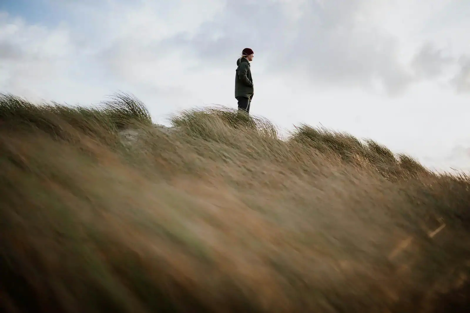 A person wearing a jacket and hat stands alone on top of a grassy hill under a cloudy sky.