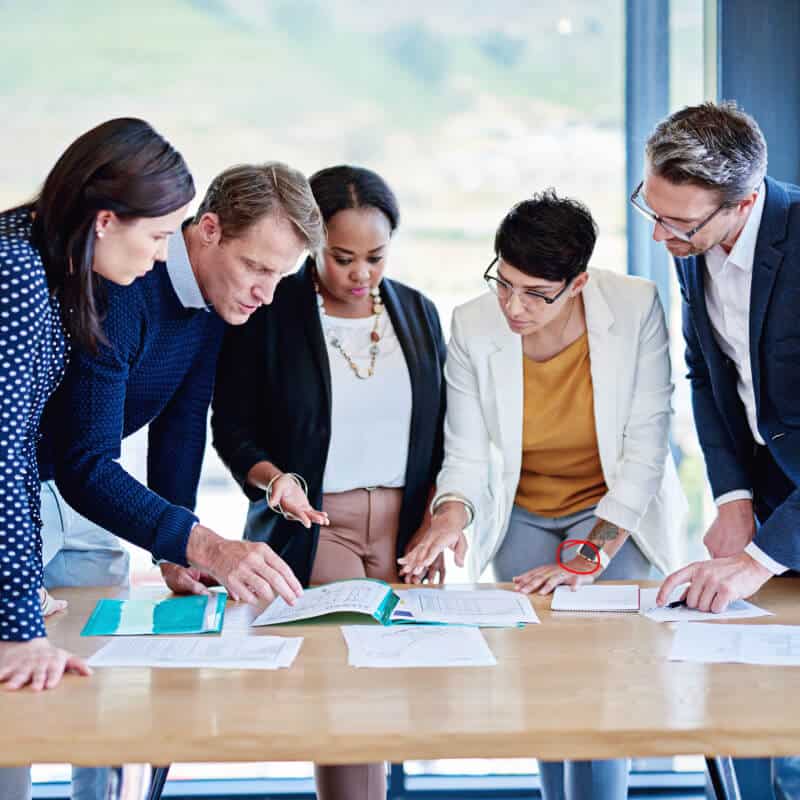 Five people stand around a table in an office, closely reviewing and discussing documents spread out in front of them.