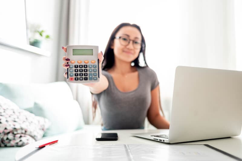 A woman sitting at a desk with a laptop holds a calculator toward the camera, with papers, a pen, and a smartphone on the table.