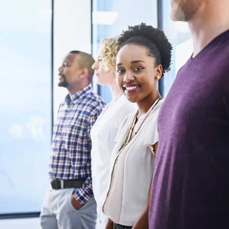 Four people stand in a row indoors; the woman in front is smiling at the camera while the others look forward. They are casually dressed and standing by large windows.