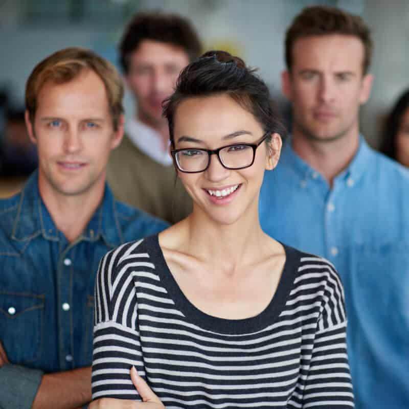 Four adults stand together indoors; a woman in glasses and a striped shirt is in focus at the front, with three men in casual clothing blurred in the background.