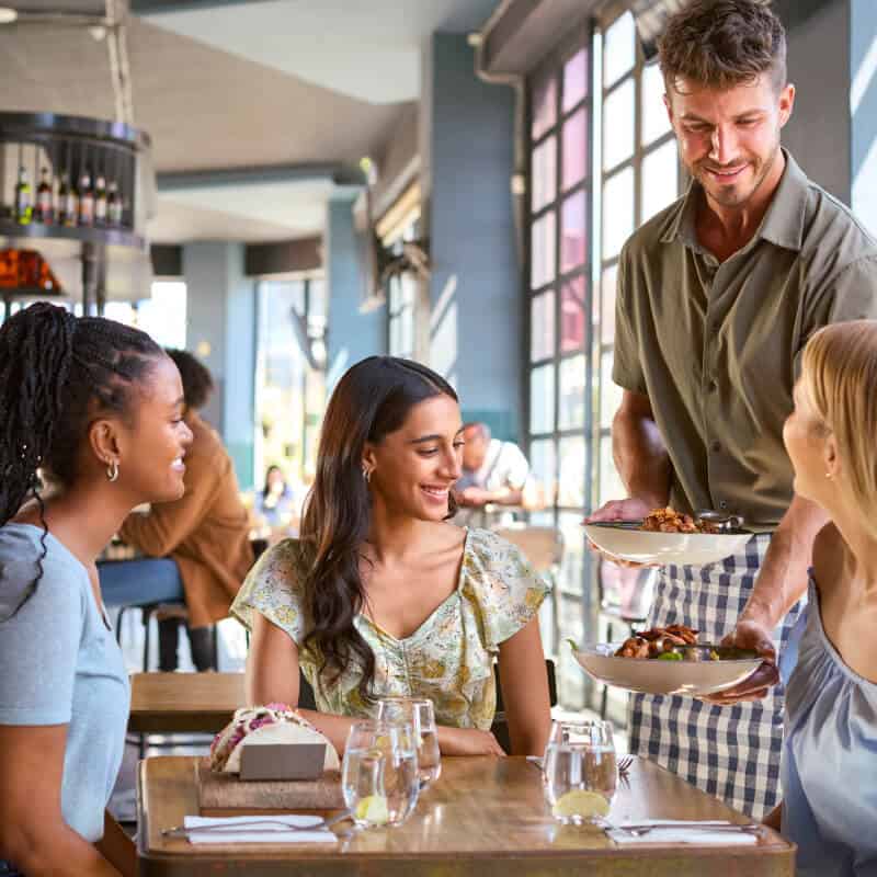 A waiter serves food to three women sitting at a restaurant table with drinks, plates, and cutlery, while natural light enters through large windows.