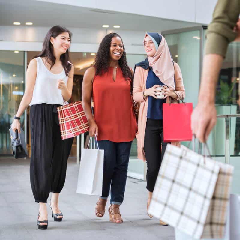 Three women holding shopping bags walk and smile outside a shopping center. Another person carrying bags is partially visible in the foreground.