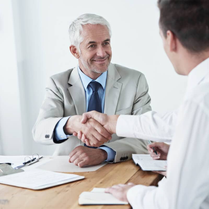 Two men in business attire shake hands across a desk in an office setting, with documents and a pen visible on the table.