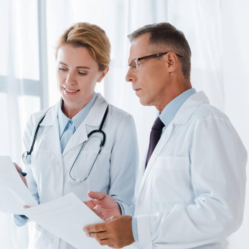 Two doctors in white coats review documents together in a bright medical office.