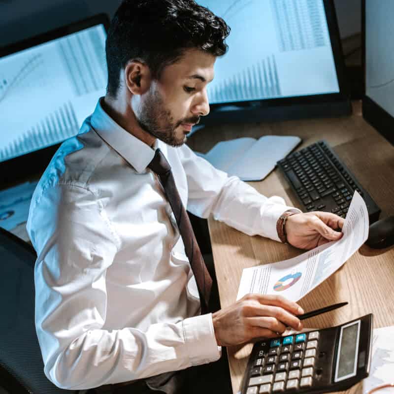 A man in a white shirt and tie reviews printed charts at a desk with a calculator, keyboard, and computer monitors displaying graphs.