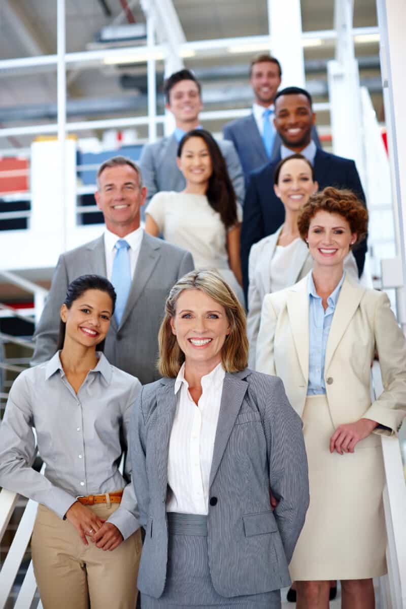 A group of nine professionally dressed people stand on stairs in an office building, smiling at the camera.