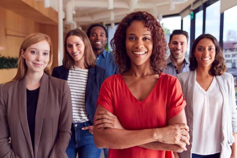 A group of six professionally dressed people stand together in an office, with a woman in a red shirt confidently posing at the front.