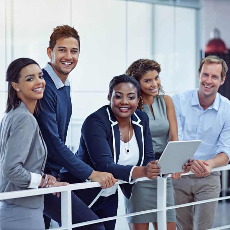 Five professionally dressed people stand by a railing in an office, smiling at the camera; one woman holds a tablet.