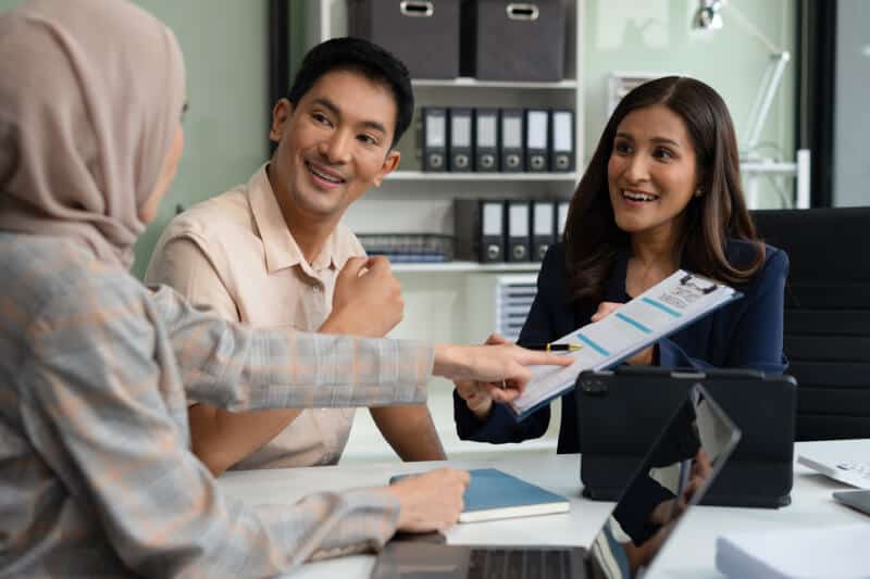 Three people sit at a desk in an office, discussing documents and smiling; one person points at a clipboard with papers.
