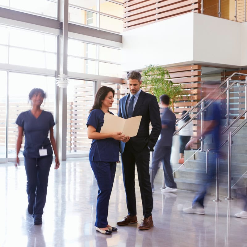 A woman in scrubs shows a folder to a man in a suit in a modern hospital lobby as other people walk by.