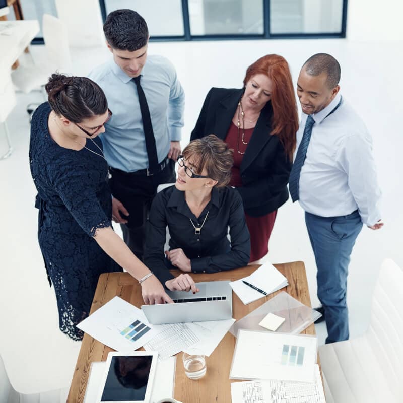 Five professionals gather around a desk with documents and a laptop, engaged in discussion in a modern office setting.
