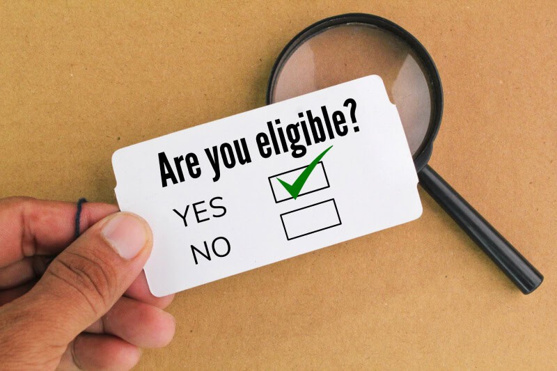 A hand holds a card labeled "Are you eligible?" with checkboxes for yes and no; the yes box is checked. A magnifying glass lies on a brown surface in the background.