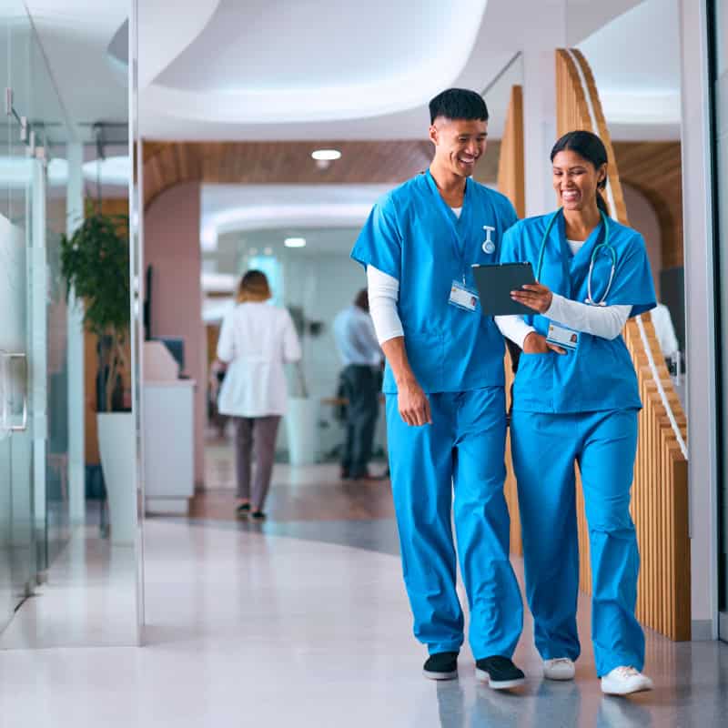Two healthcare professionals in blue scrubs walk down a hospital hallway, looking at a digital tablet and smiling.