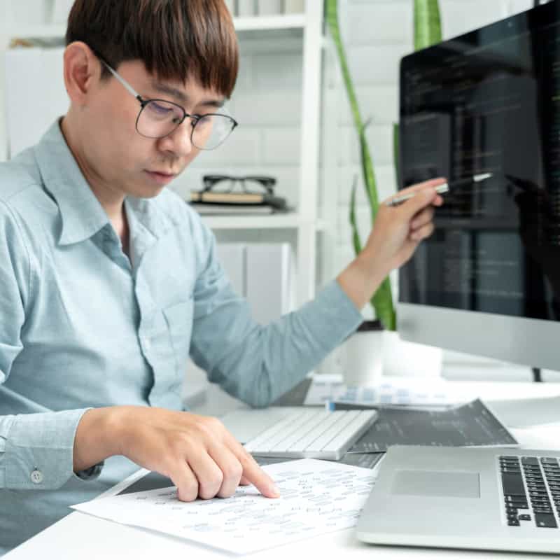 A person in glasses works at a desk, pointing at a printed diagram and computer code on a monitor, with a laptop and plants nearby.