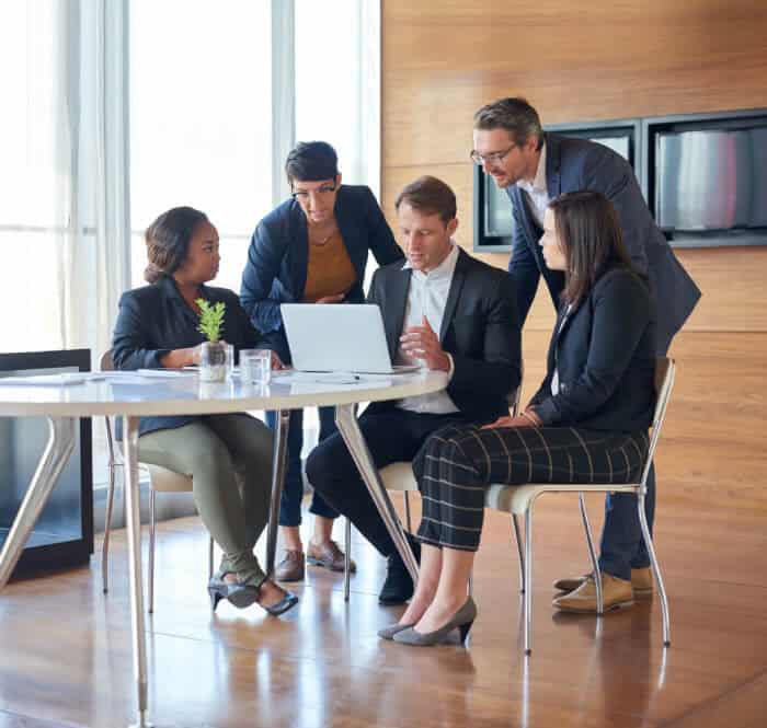 Five people in business attire gather around a table, looking at a laptop screen during a meeting in a modern office setting.