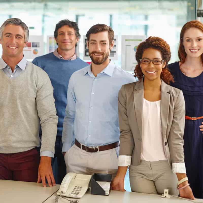 A group of five professionally dressed people stand together in an office setting, smiling at the camera. A desk with a phone and papers is in the foreground.