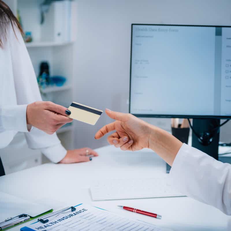 A person hands a card to a healthcare professional in an office, with a health insurance form and pens on the desk and a computer screen in the background.
