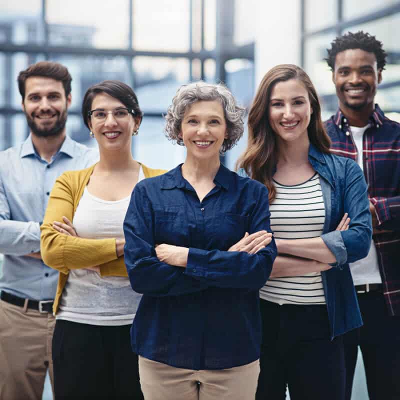 Five people stand indoors with arms crossed, smiling at the camera; the group is diverse in age, gender, and ethnicity, and they are dressed in casual business attire.