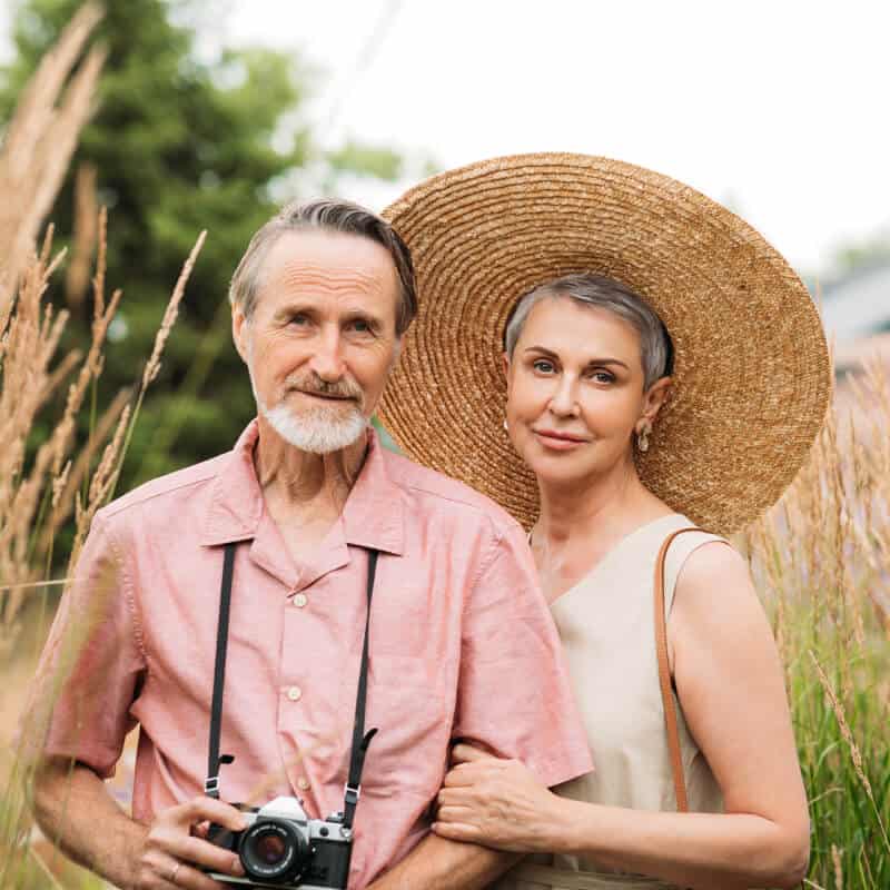 An older man holding a camera and an older woman wearing a large straw hat stand together outdoors in tall grass.