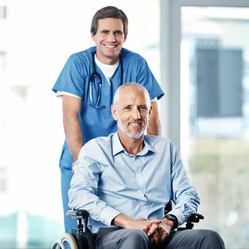 A healthcare professional in scrubs stands behind an older man seated in a wheelchair, both smiling in a bright indoor setting—reflecting compassionate care and personalized Medicare consulting.