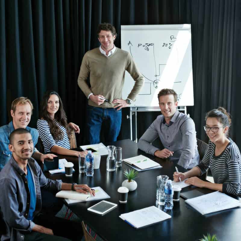 Six people sit around a table with notebooks and water bottles, while one person stands by a whiteboard with graphs and diagrams drawn on it.